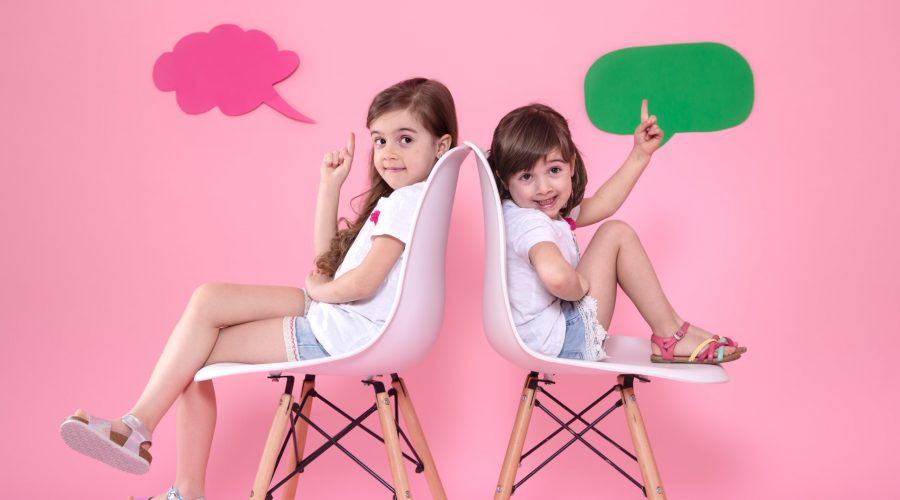 Two little girls on a colored pink background are sitting on chairs with speech icons . The concept of communication and friendship